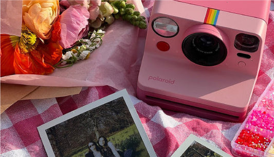 Pink Polaroid instant camera surrounded by flowers with printed instant photos on a picnic blanket